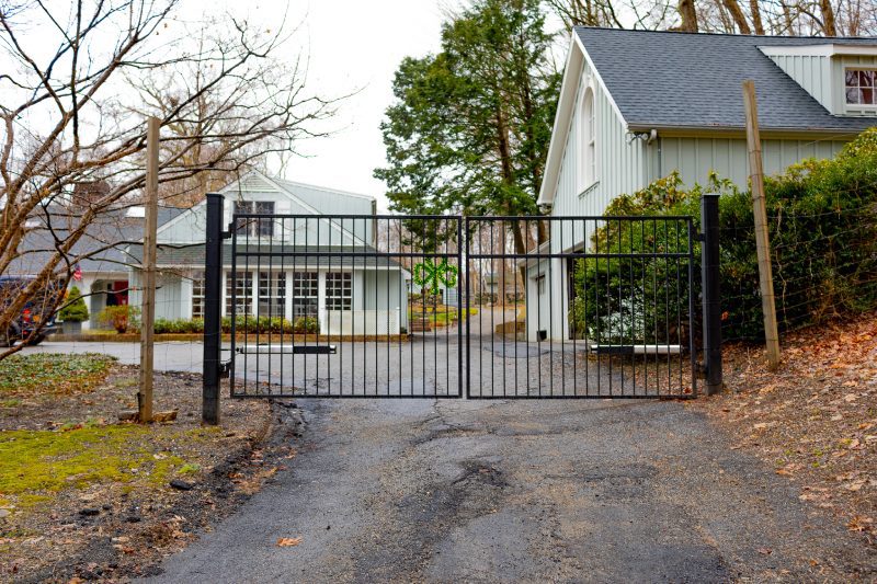 A Black Metal Gate with Thin Pickets Stands in Front of a Farmhouse Style Home.
