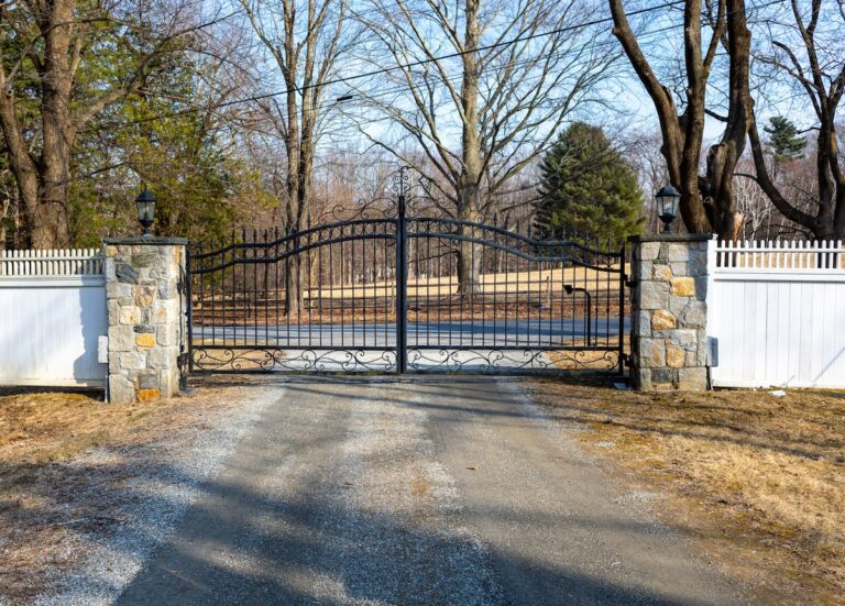 iron swing driveway gate with swirl design and stone columns with white wood fencing