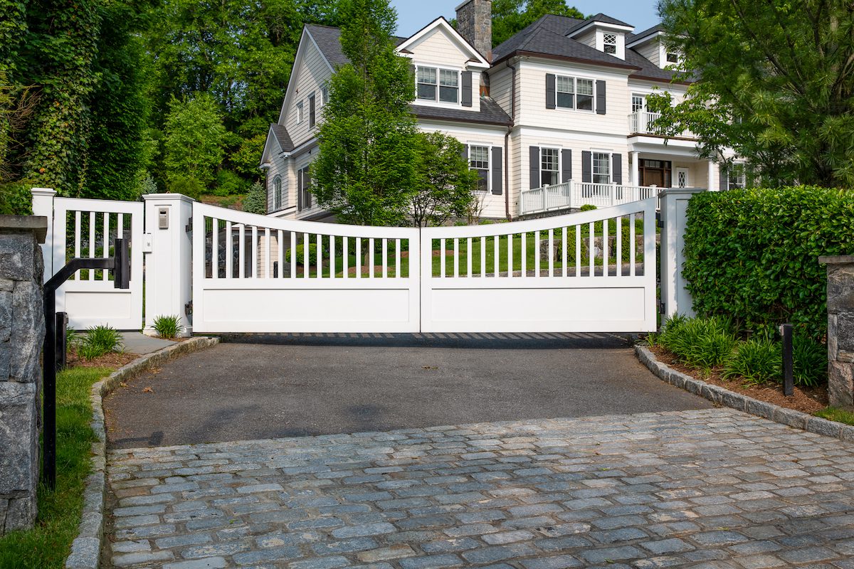 A white composite automatic driveway gate guards a gently sloping driveway leading up a hill to a large Colonial Revival house.