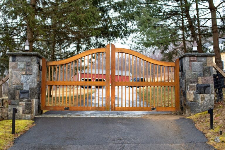 Arched farmhouse wood driveway gate with lots of spacing between slats and natural stone columns with lighting